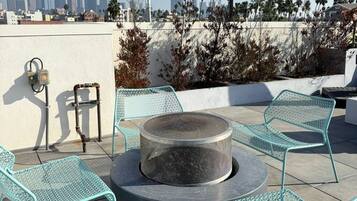 A rooftop patio with a round fire pit, metal chairs, and a city skyline view.