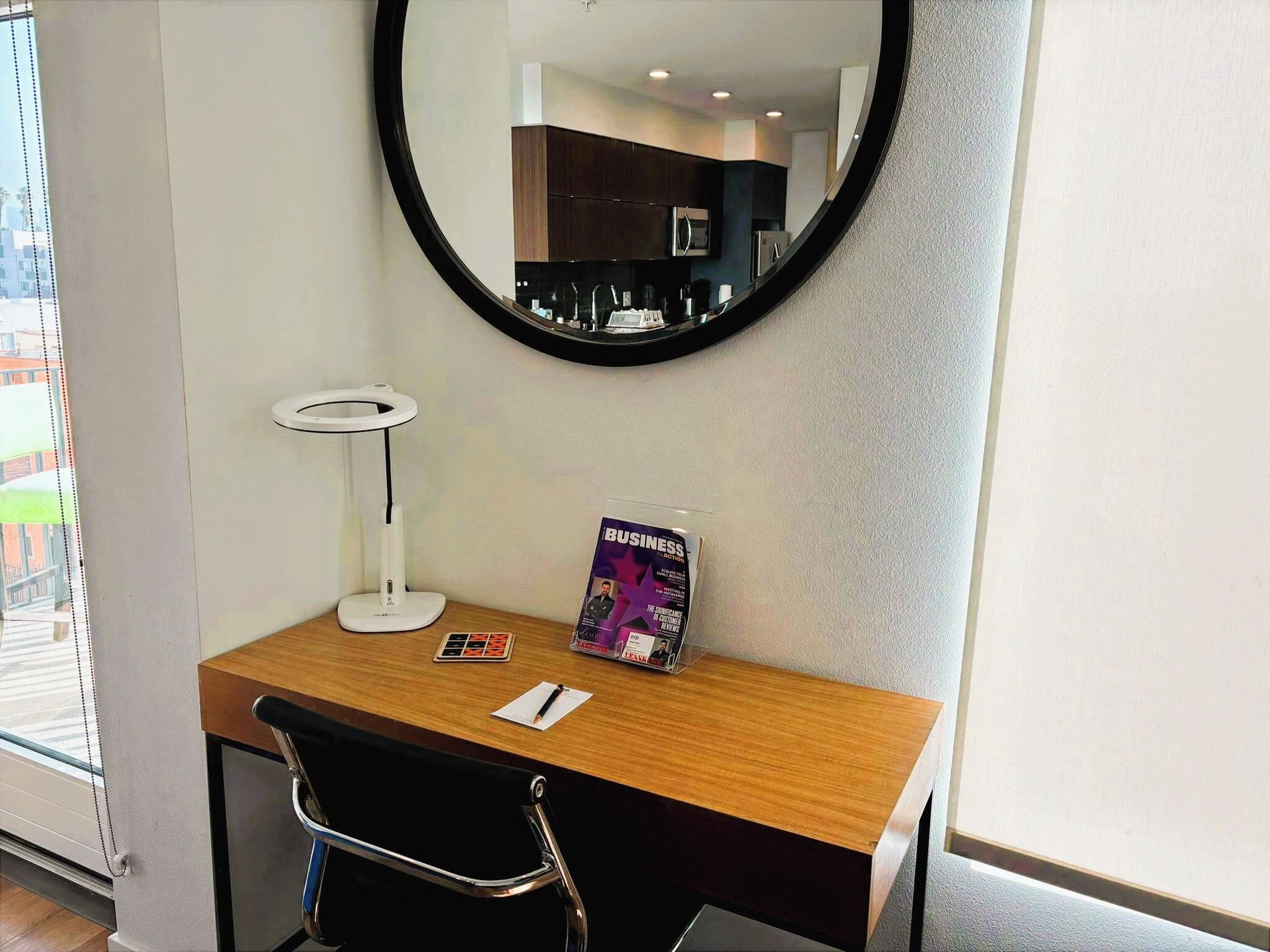Ein Hotelzimmer mit einem Holztisch, einem schwarzen Stuhl, einer weißen Schreibtischlampe, einem Spiegel und einer Zeitschrift auf dem Tisch.