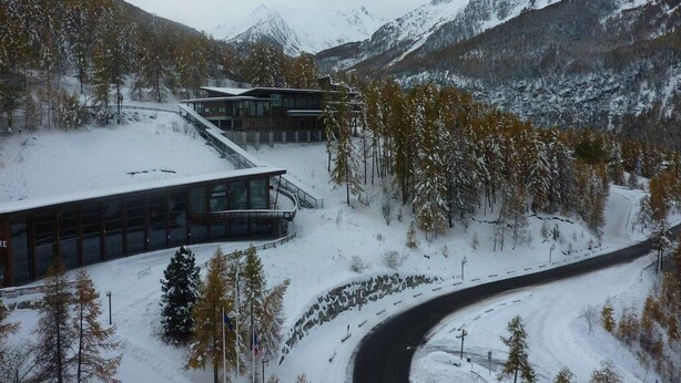Un bâtiment moderne doté de grandes fenêtres et d’une allée incurvée, entouré d’arbres et de montagnes couverts de neige.