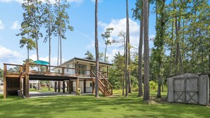 A two-story house with a covered patio and a garage, surrounded by tall trees and a well-maintained lawn. - Waterfront 2BR/1BA – Fully Furnished Home in Covington, LA (Covington)