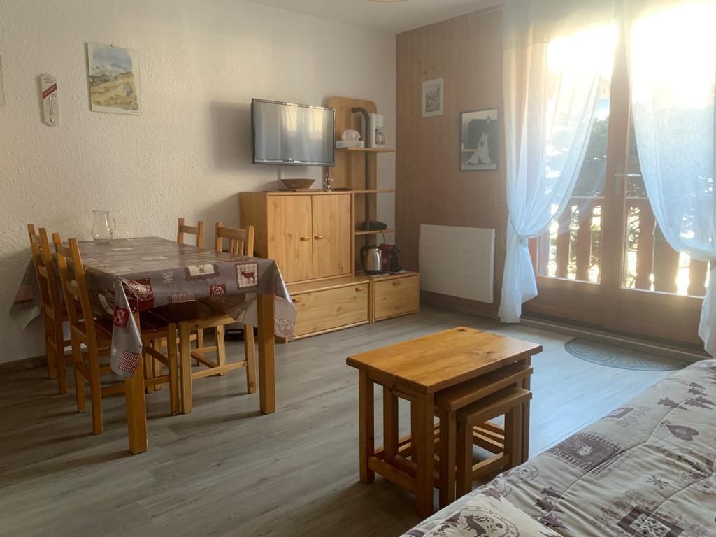 Een gezellige kamer met een houten eettafel, stoelen, een klein houten krukje, een tv op een houten dressoir en een bed met beddengoed met een patroon.