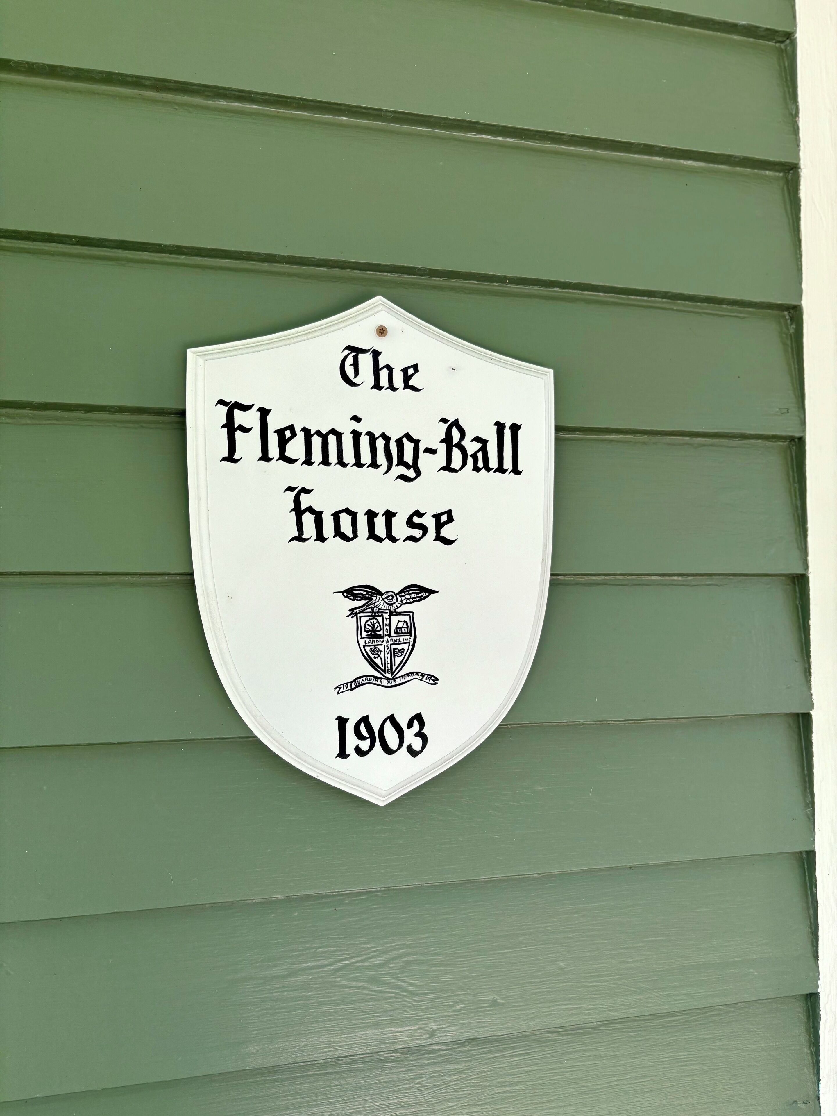 A white shield-shaped sign with black lettering stating 'The Fleming-Ball House 1903' on a green wooden wall.