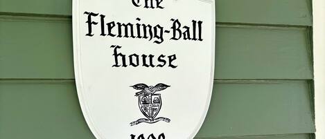 A white shield-shaped sign with black lettering stating 'The Fleming-Ball House 1903' on a green wooden wall.