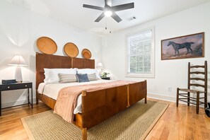 Un dormitorio con una cama de madera, una mesita de noche, un escritorio, una silla, un ventilador de techo, una ventana con persianas y un cuadro en la pared.