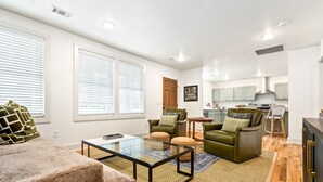 A modern living room with a glass coffee table, a sofa, and a green armchair. There is a kitchen in the background with light green cabinets and a stainless steel hood. - Gorgeous Auburn Getaway! BRAND NEW 3Bed/3Bath (Auburn)