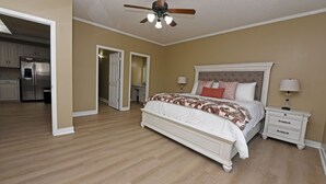 A bedroom with a large bed, a nightstand, a ceiling fan, and a view into the kitchen.