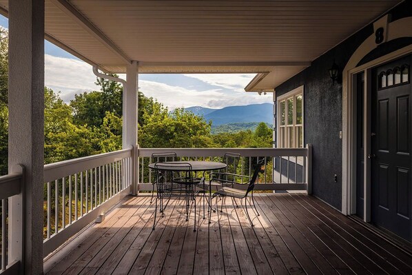 Une terrasse en bois avec une table et des chaises, une balustrade blanche et une vue sur la verdure et les montagnes.