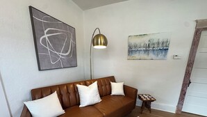 A brown leather sofa with white cushions, a round rug, a framed picture, a floor lamp, and a small wooden table with a remote control.