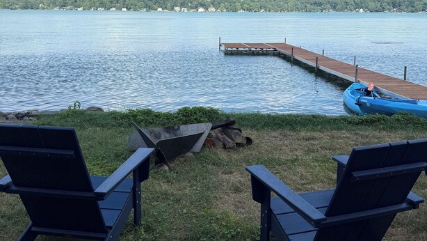 A lakeside view with a wooden dock extending into the water, two black lounge chairs facing the lake, and a blue kayak on the dock.