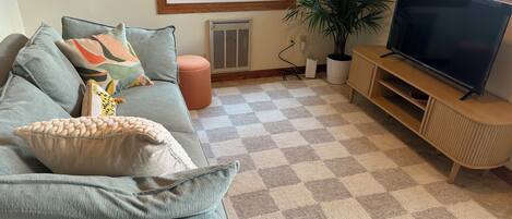 A living room with a checkered carpet, a grey sofa with multiple cushions, a television on a wooden stand, and a potted plant.