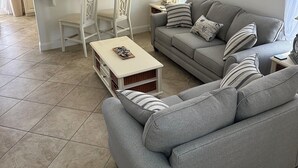 A modern living room with a grey sofa, a small white table, and a kitchen area with light-colored cabinets and a black refrigerator. - Southern Dunes- Lake View Villa -Private Pool (Haines City)