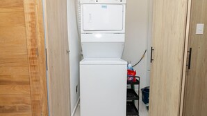 A compact laundry room with a stacked washer and dryer, a blue bucket, and a red bag on a shelf.