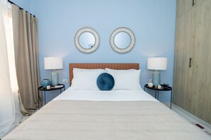 Una cama bien hecha con sábanas blancas y un cojín azul decorativo, flanqueada por dos mesitas de noche con lámparas y adornos.
