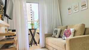 Un saló amb un sofà beix, una televisió sobre un suport de fusta, un ventilador de sostre i una vista d'un edifici a través d'una finestra.