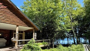 A wooden cabin with a covered porch, a small garden, and a view of a lake surrounded by trees. - Welcoming 4-bedroom cabin in Chesterville (Chesterville)