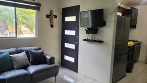 Una sala de estar con un sofá de color oscuro, una puerta negra con paneles blancos, un televisor montado y una zona de cocina con refrigerador y armarios.