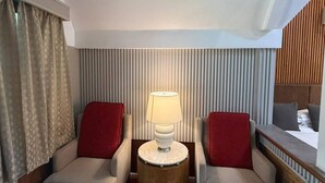 Ein Hotelzimmer mit zwei Sesseln, einem kleinen runden Tisch mit Lampe, einem Bett mit Holz Kopfteil und einem Fenster mit Vorhängen.