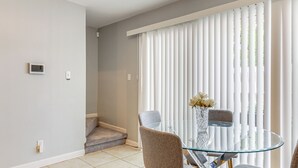 Un comedor con una mesa de vidrio y cuatro sillas, un florero con flores, y vista a través de persianas verticales.