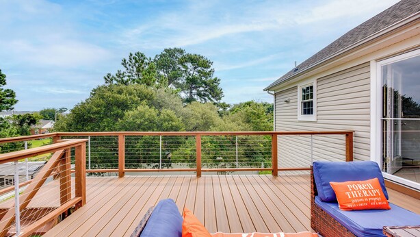 Una terraza de madera con zona de estar acolchada, un ventanal de vidrio que da a una habitación y una vista de árboles y una casa al fondo.