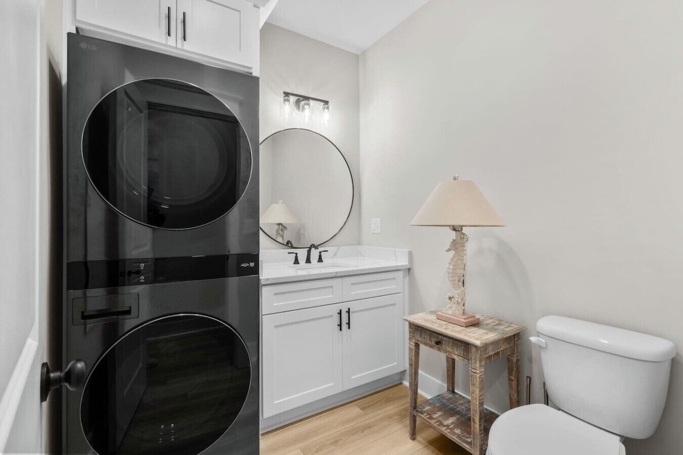 A compact bathroom with a modern black washing machine, a white sink with a mirror above it, a small wooden side table with a lamp, and a white toilet.