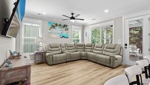 A modern living room with a sectional sofa, a ceiling fan, and a view through a glass door. - Luxury Beach House, Elevator, Golf Cart & Grill! (Myrtle Beach)