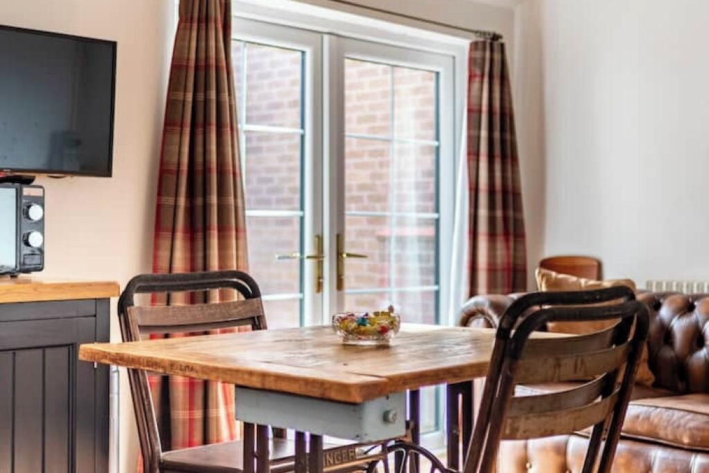 A dining area with a wooden table, two chairs, a television, and a window with plaid curtains.