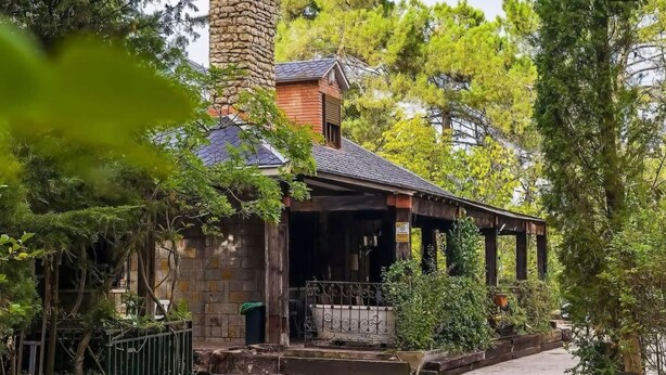 A rustic building with a stone chimney, a porch with columns, and lush greenery.