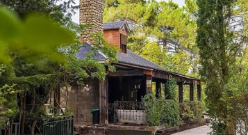 A rustic building with a stone chimney, a porch with columns, and lush greenery.