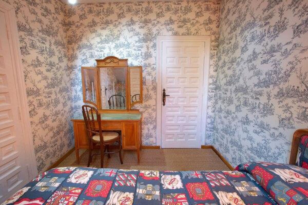 Ein Schlafzimmer mit floraler Tapete, einem hölzernen Schreibtisch mit Stuhl und einem Bett mit gemusterten Bettbezügen.