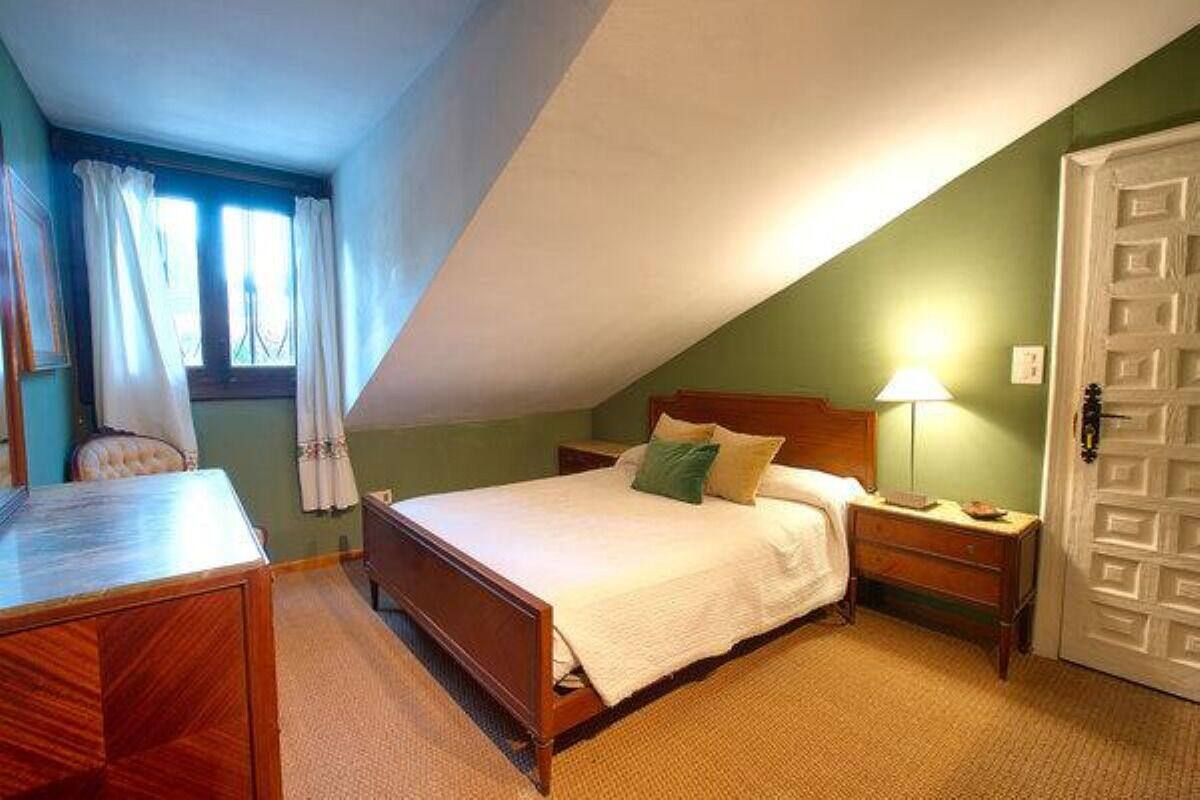 A bedroom with a wooden bed, a nightstand, a desk, and a chair. There is a window with curtains, a door, and a lamp on the nightstand.
