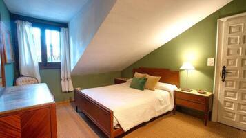 A bedroom with a wooden bed, a nightstand, a desk, and a chair. There is a window with curtains, a door, and a lamp on the nightstand.