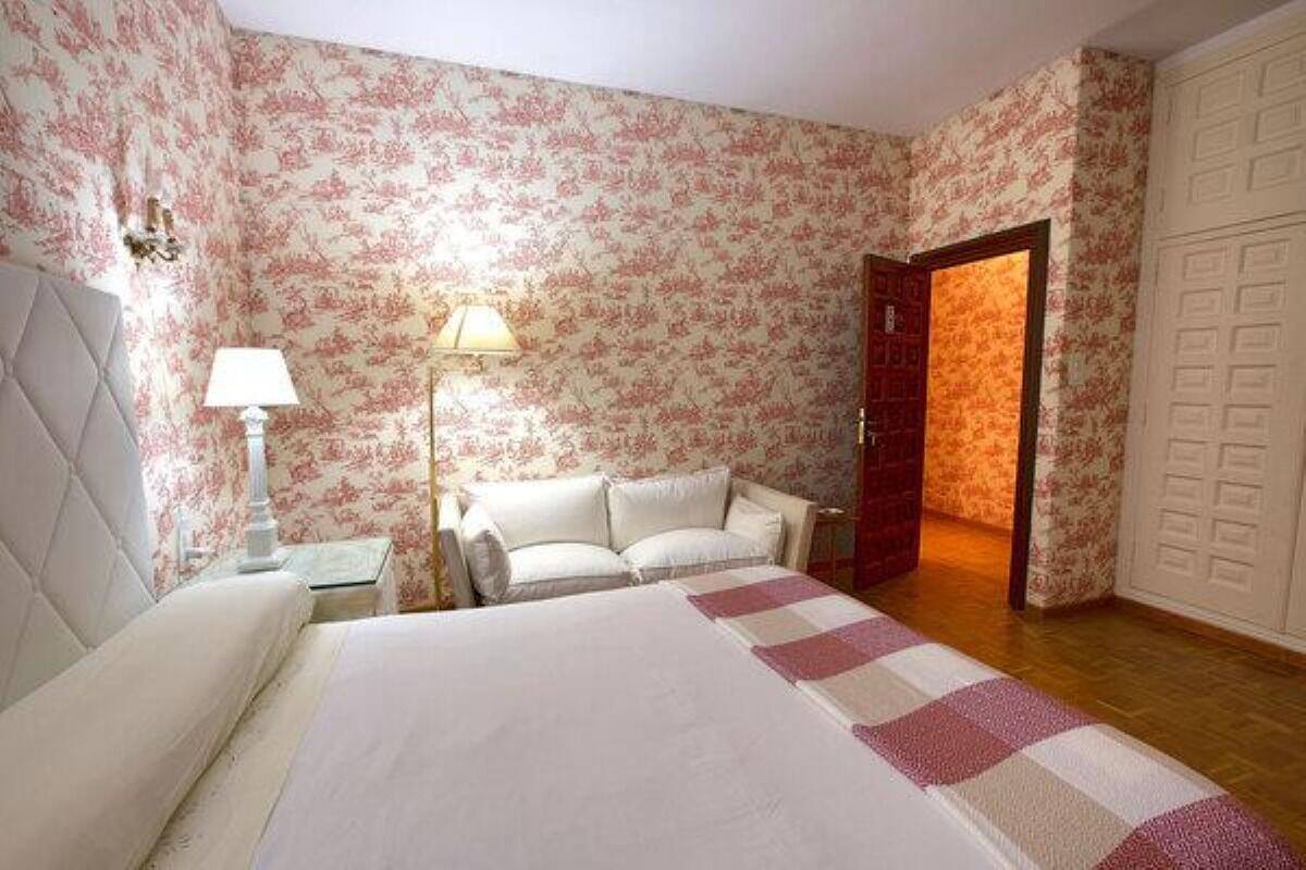 A bedroom with floral wallpaper, a bed with a checkered bedspread, a nightstand with a lamp, and a door leading to another room.