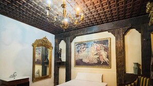 A bedroom with a wooden ceiling, a chandelier, a bed, a chair, and a painting on the wall.