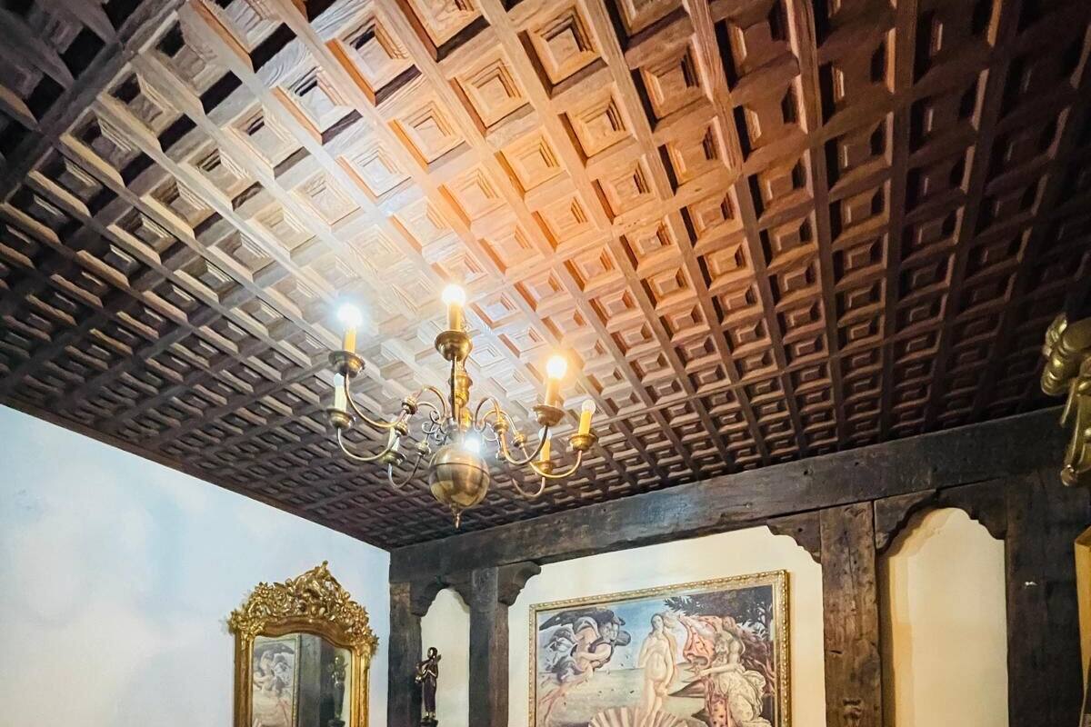 A bedroom with a wooden ceiling, a chandelier, a bed, a chair, and a painting on the wall.