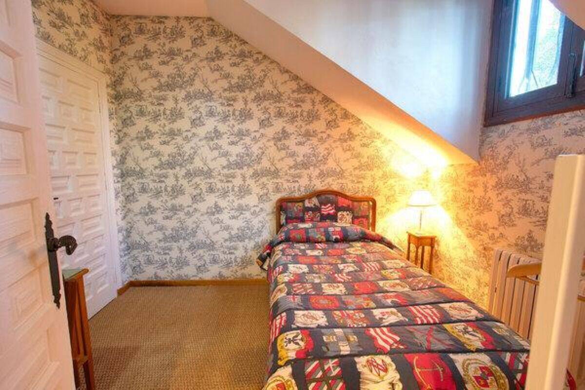 A bedroom with a sloped ceiling, patterned wallpaper, a bed with a quilt, a nightstand, and a window with natural light.