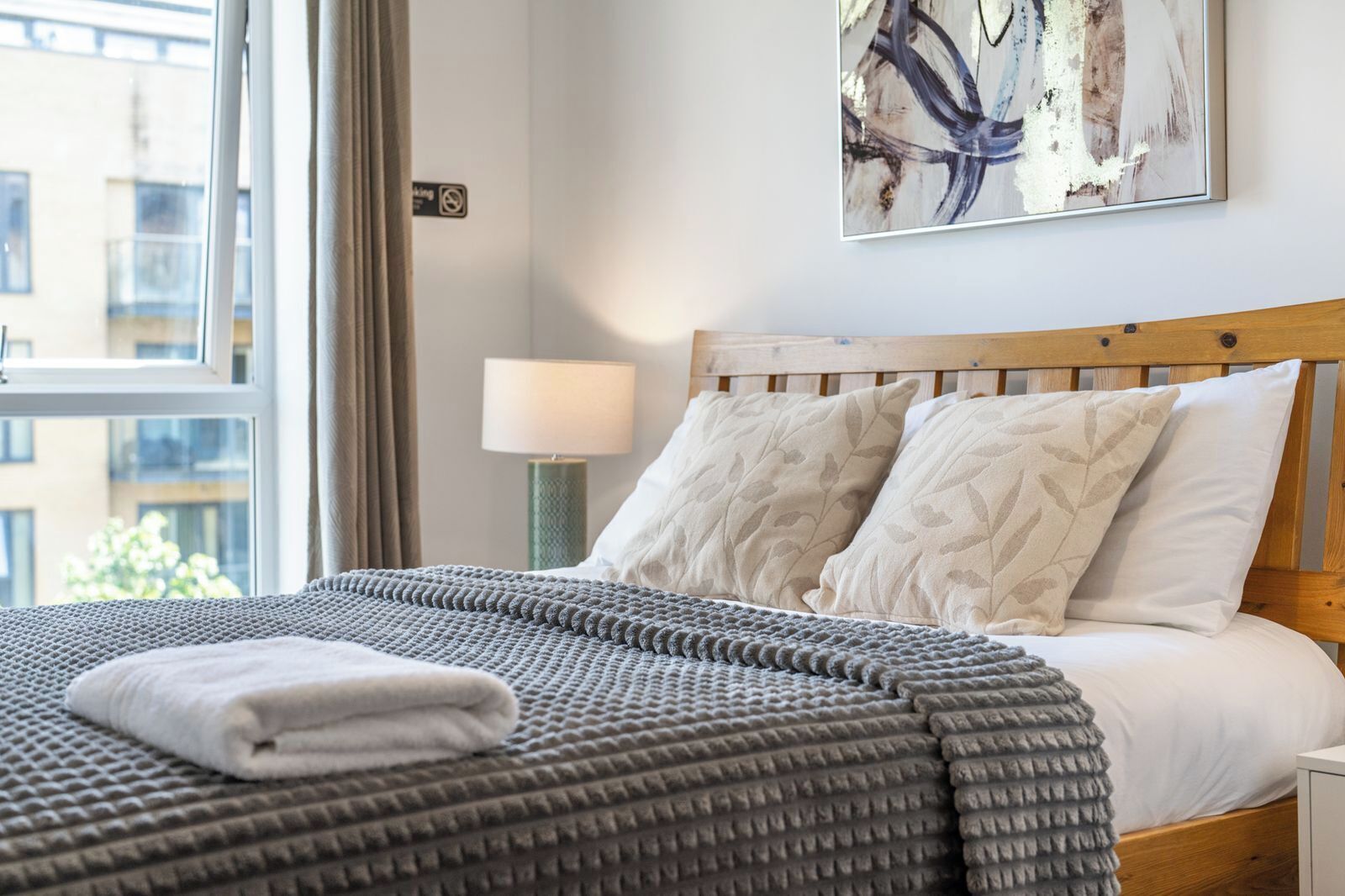 A neatly made bed with a grey textured blanket and white pillows, a bedside lamp, and a painting on the wall.