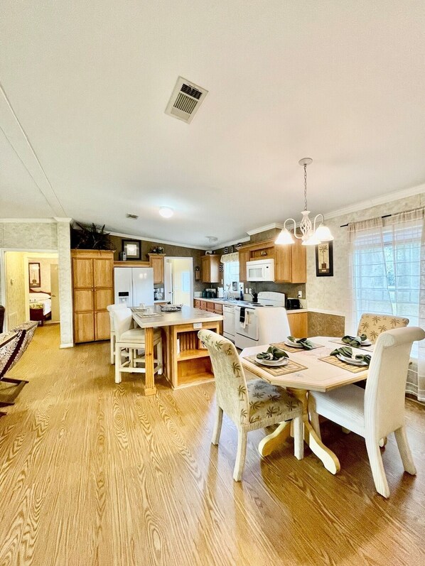 Un comedor amplio con una mesa y sillas de madera, una cocina al fondo y un ventanal con cortinas.