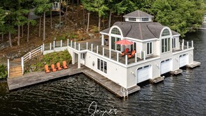 Una casa flotante blanca con cubierta y una sombrilla roja, amarrada en un lago tranquilo rodeado de árboles.
