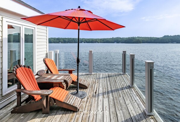 A wooden deck with two orange Adirondack chairs and a red umbrella, overlooking a calm lake with a forested shoreline.