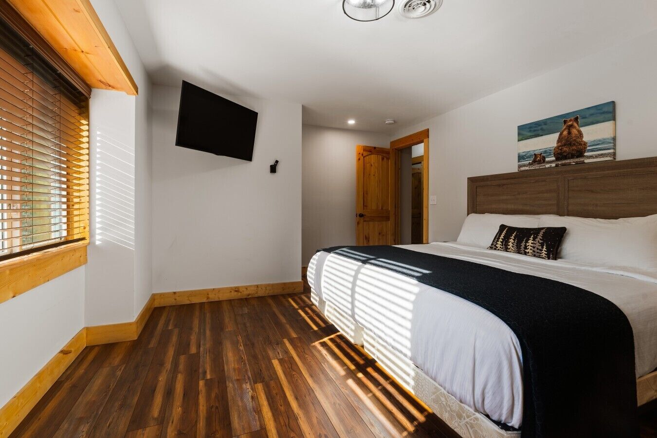 Un dormitorio con piso de madera, una cama con cubrecama blanco y negro, un televisor montado y un cuadro en la pared.