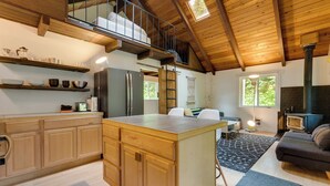 A spacious kitchen with wooden cabinets, a central island, and a loft bedroom above. - Tiny: An A-Frame Cabin in the Woods (Rhododendron)