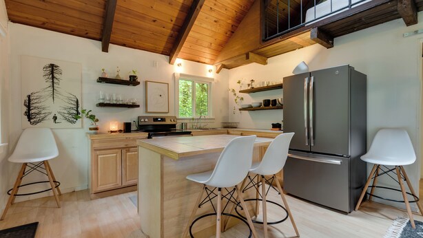 A modern kitchen with wooden cabinets, a large island with chairs, a stainless steel refrigerator, and a window providing natural light. - Tiny: An A-Frame Cabin in the Woods (Rhododendron)