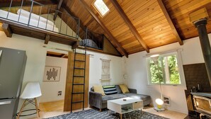 A cozy loft-style room with a loft bed, a sofa with cushions, a white coffee table, a fireplace, and a large window. - Tiny: An A-Frame Cabin in the Woods (Rhododendron)