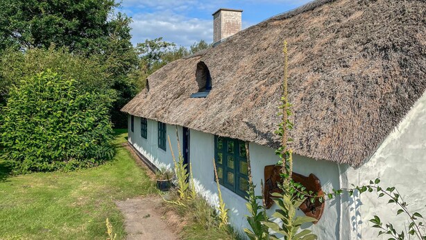 Ein traditionelles, mit Stroh gedecktes Haus, verbunden mit einem schmalen Pfad, umgeben von viel Grün.