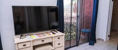 A room with a large flat-screen TV on a white dresser, a small table with a blue chair, and a sliding glass door with blue curtains.