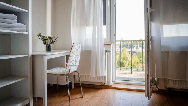 A room with a wooden floor, a white desk with a chair, a radiator, and a view of a balcony with a railing.