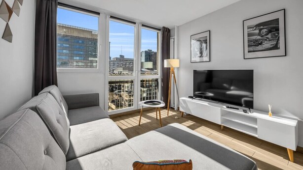 A modern living room with a grey sofa, a flat-screen TV, and a city view through large windows. - Rondo ONZ Apt - 11th Floor, Top location, Metro - by Rentujemy (Warszawa)