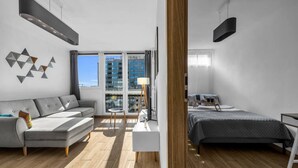 A modern, well-lit room with a grey sofa, a bed, and a desk. Large windows provide natural light and a view of the city. - Rondo ONZ Apt - 11th Floor, Top location, Metro - by Rentujemy (Warszawa)