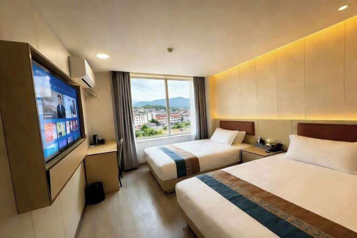 Habitación de hotel con dos camas, televisión, ventana y vista a la ciudad, además de un escritorio con lámpara.