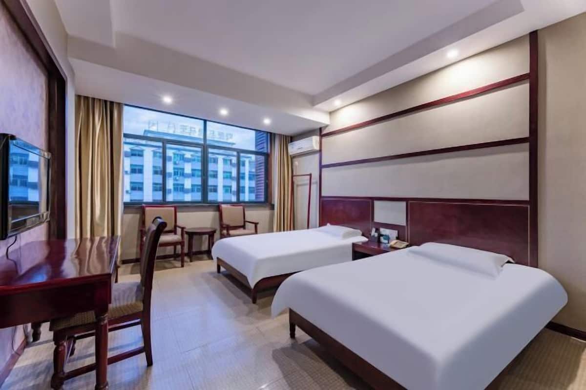 Ein Hotelzimmer mit zwei Betten, einem Schreibtisch und einem Stuhl. Ein großes Fenster bietet einen Blick auf Gebäude außerhalb.